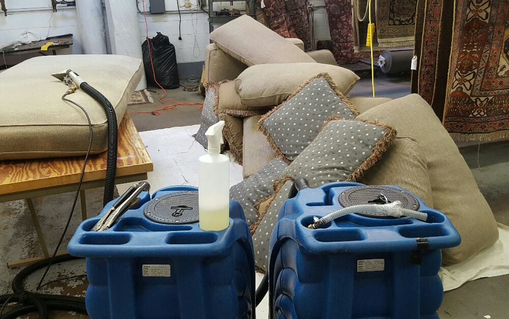 Pile of sofa cushions laying on white tarp. Two large blue upholstery steamer machines are prepped and ready to clean.