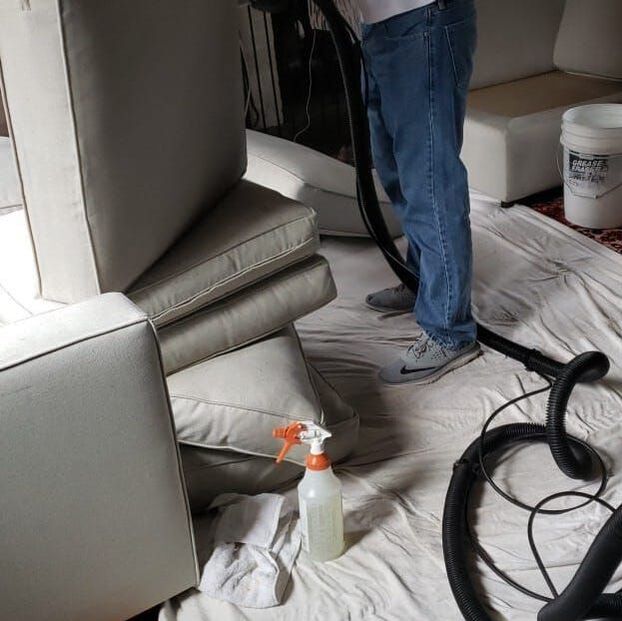 Contractor cleaning gray sofa cushions on top of white tarp. Spray bottle of upholstery cleaner and steam cleaner being used by contractor.