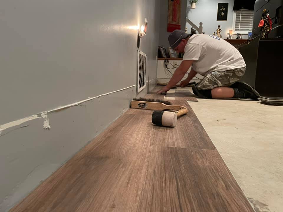 Contractor installing wood like vinyl flooring in gray room. Handle held brush broom and mallet laying on flooring.