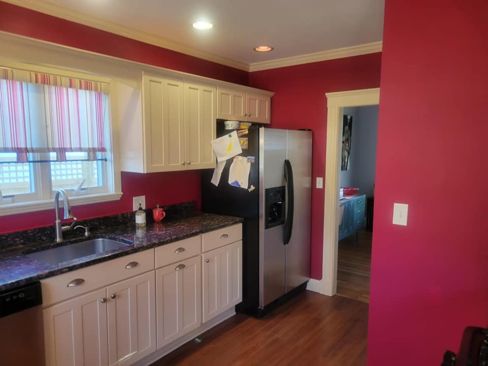 Kitchen with kitchen sink and window view. White cabinets, black countertops and stainless steel refrigerator and newly painted red walls with white trim.