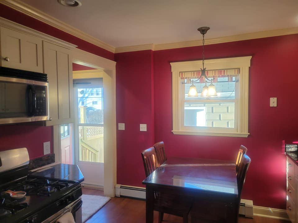 Stove, microwave and dining room table view kitchen. Window above dining table and front door. Newly painted red walls against white trim.