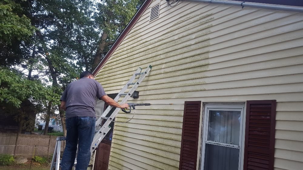 Contractor power washing dirty siding of beige house with brown shutters