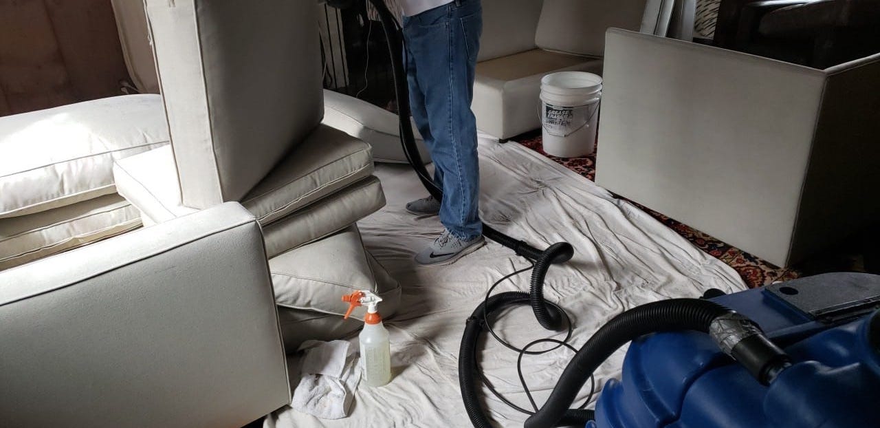 Contractor cleaning gray sofa cushions on top of white tarp. Spray bottle of upholstery cleaner and steam cleaner being used by contractor.